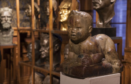 Fragment pomieszczenia wypełnionego rzeźbami Alfonsa Karnego. Są to głowy wykonane w różnych materiałach, na pierwszym planie rzeźba raczkującego dziecka.