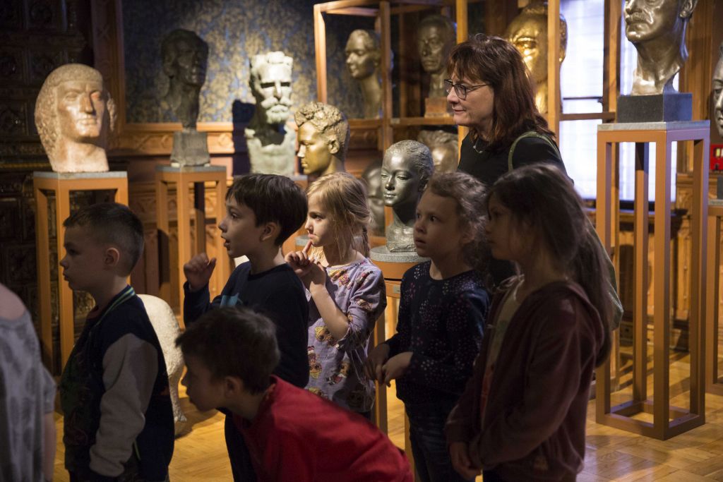Pani z grupką sześciorga dzieci stoi w sali z brązowymi ścianami i tapetą. W tle rzeźby Alfonsa Karnego.