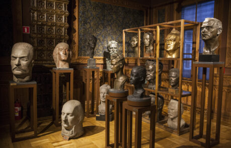 Wnętrze dużego pomieszczenia z brązowymi ścianami, tapetami i piecem. W pomieszczeniu stoją rzeźby Alfonsa Karnego. Przedstawiają głowy znanych Polaków, na przykład Romualda Traugutta, Mikołaja Kopernika, Władysława Broniewskiego.