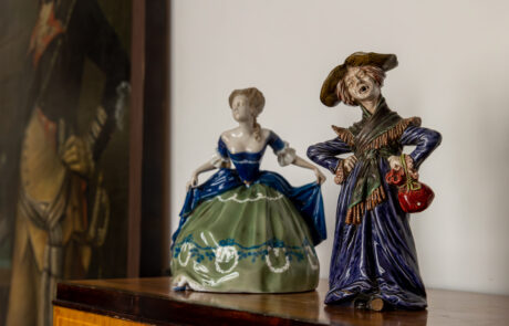 Dwie porcelanowe figurki kobiet stoją na brązowej drewnianej komodzie. Z lewej strony młoda dama w balowej sukni, z prawej staruszka w niebieskiej sukni i kapeluszu. W tle obraz żołnierza.