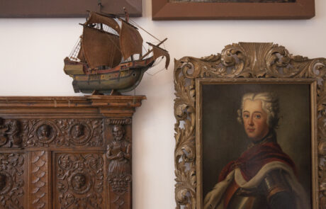 Dzieła sztuki z kolekcji Alfonsa Karnego. Z lewej strony drewniany mebel z płaskorzeźbami i stojącym na nim modelem żaglowca, po prawej w ozdobnej ramie portret młodego mężczyzny w zbroi i peruce.