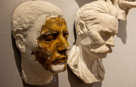 Na ścianie wiszą dwie maski pośmiertne wykonane z gipsu. Maska z lewej strony częściowo pomalowana jest na brązowy kolor.