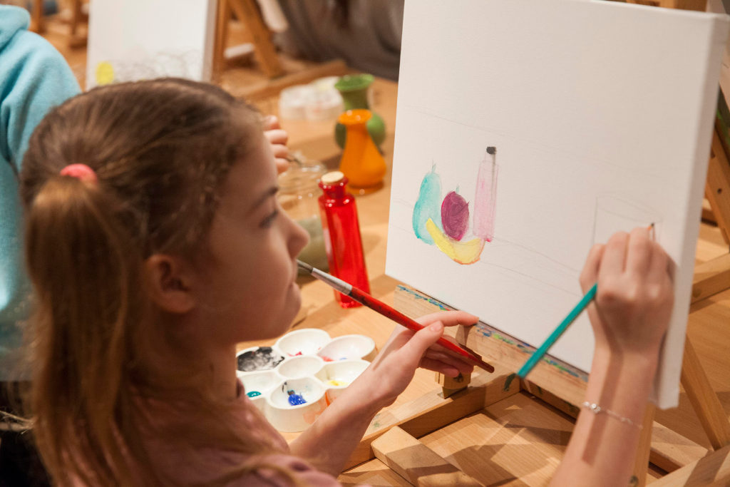 Kilkuletnia dziewczynka trzyma w prawej dłoni ołówek, w lewej pędzel i rysuje obraz na postawionej przed nią małej sztaludze. Obok siebie na palecie ma farby