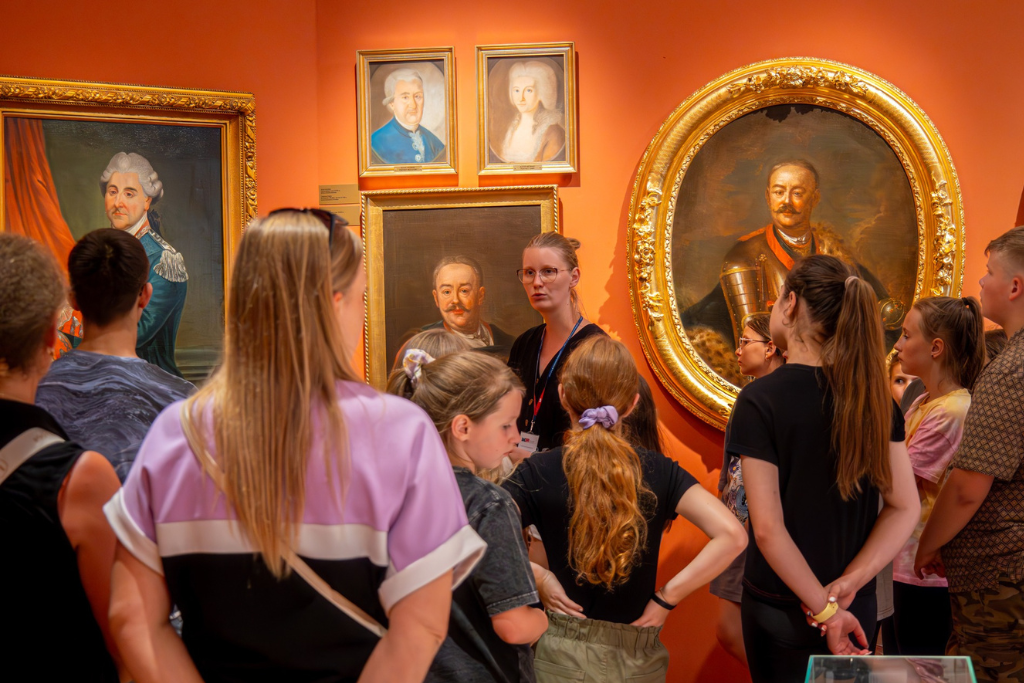Przewodnik muzealny oprowadza grupę zwiedzających po wystawie stałej na Sali Czerwonej, na tle portretów z XVIII wieku.
