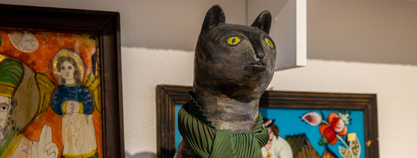 Na pierwszym planie figurka szarego kota z przewiązanym na szyi szalikiem z zielonego materiału. W tle obrazy ze sceną religijną i sceną góralską.