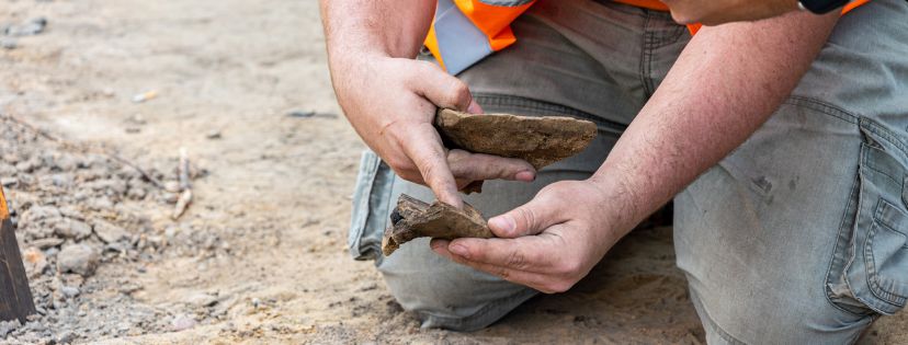 Archeolog w stroju roboczym klęczy na dnie wykopu, trzyma w dłoniach fragmenty kafla.