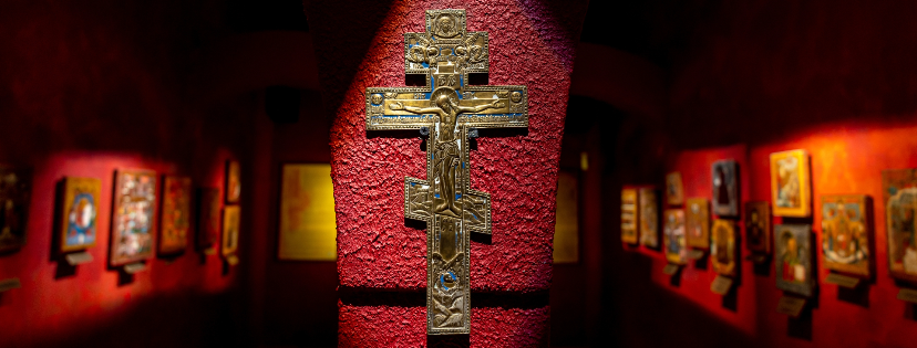 Na pierwszym planie mosiężny, emaliowany krzyż ośmiokończasty na czerwonej ścianie. W oddali w półmroku kilkanaście ikon z warsztatów staroobrzędowych Wietki, Niewiańska, Palechu.
