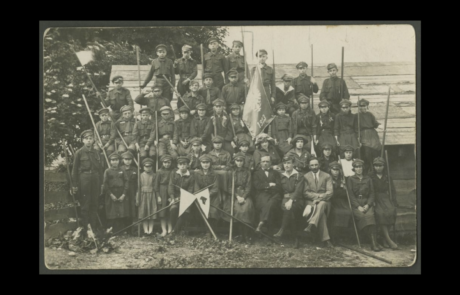 Fotografia przedstawiająca grupę młodzieży z hufca harcerskiego z 1923 r. ubrana w mundury. Przed grupą skrzyżowane drzewce proporczyków harcerskich. W tle widoczne zarośla oraz drewniany budynek.
