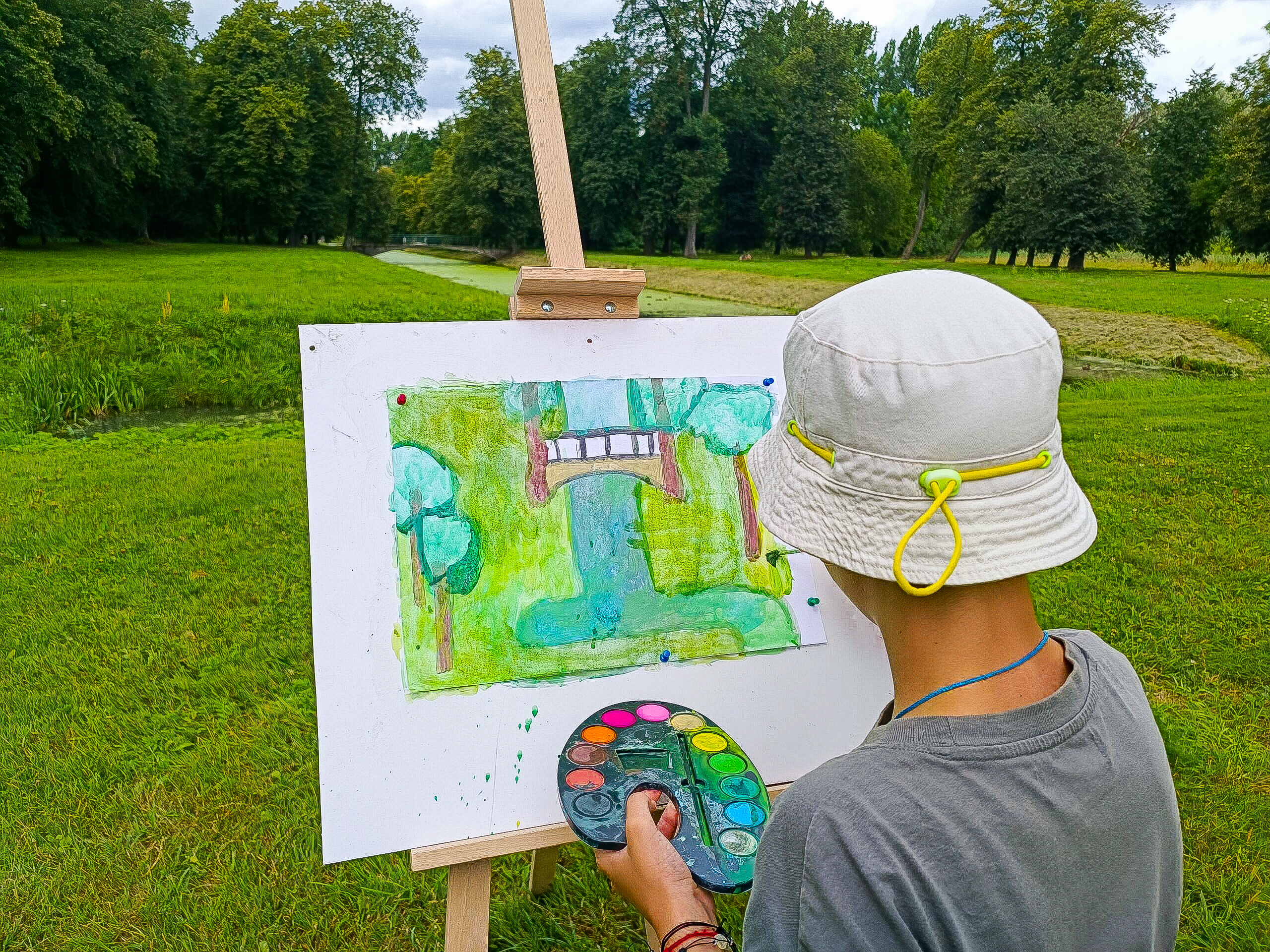 Odwrócona plecami osoba w kapeluszu stoi przed sztalugą. Na kartonie maluje widoczny przed nią fragment parku.