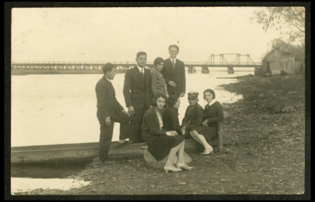 Fotografia przedstawiająca mieszkańca Tykocina Menachema Turka wraz z przyjaciółmi na tle rzeki i mostu w Tykocinie. Po prawej stronie drewniane zabudowania.