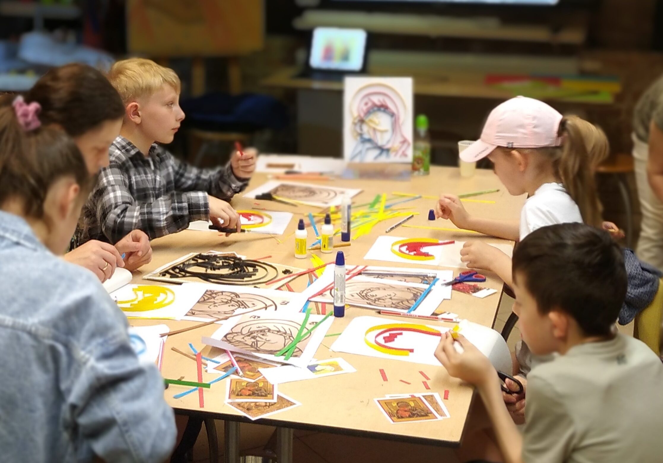 Pięcioro dzieci w wieku wczesnoszkolnym siedzi przy wspólnym stole w sali warsztatowej. Kleją kolorowymi skrawkami papieru trójwymiarowy wizerunek Matki Bożej z Dzieciątkiem „Eleusa”.